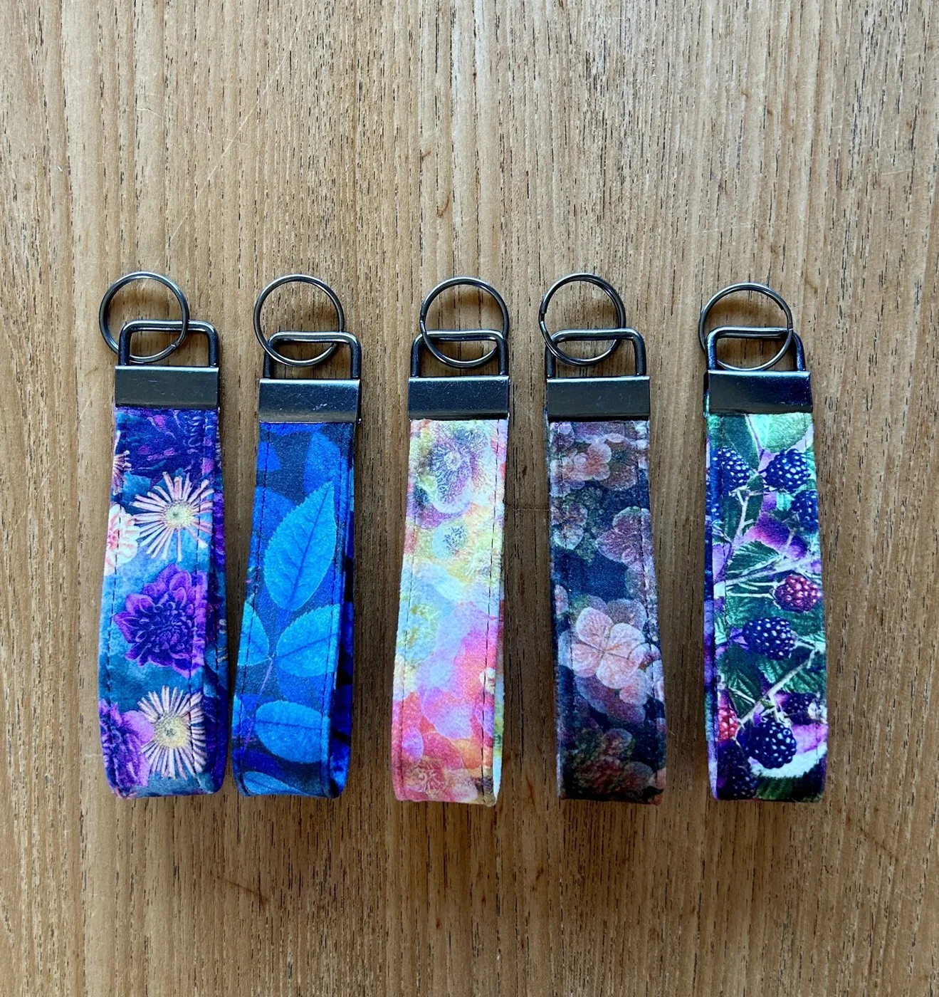 Five fabric key fobs in purple, blue and pink floral prints laid on a wooden surface
