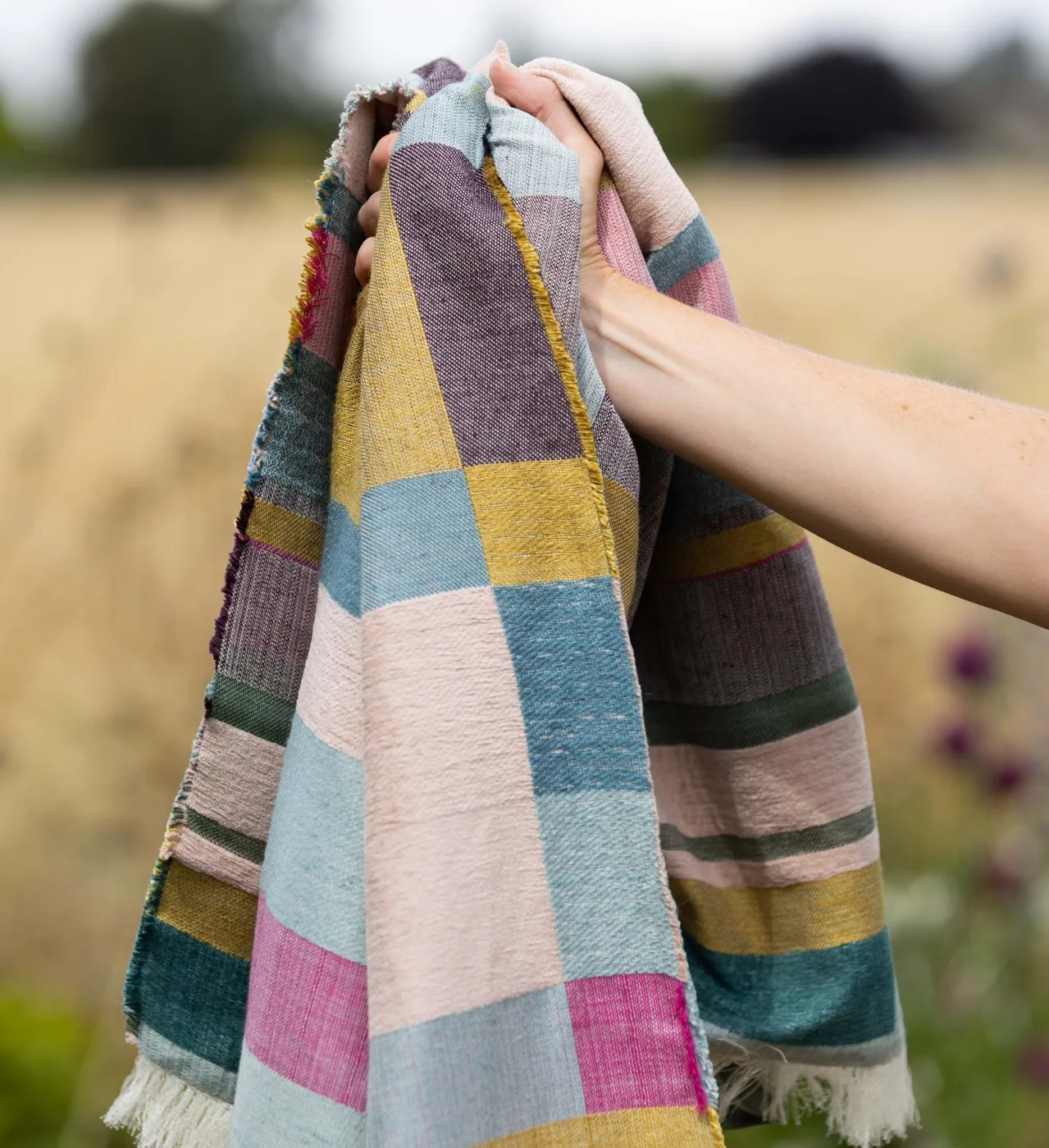 A hand holds up a woven throw in patchwork blocks of pink, green, grey and mustard against a field