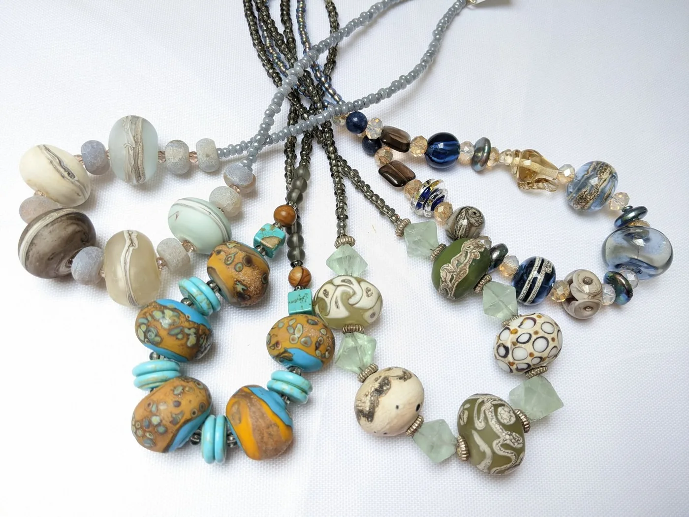 Several necklaces of large patterned glass beads in turquoise, ochre, cream and grey laid on a white surface