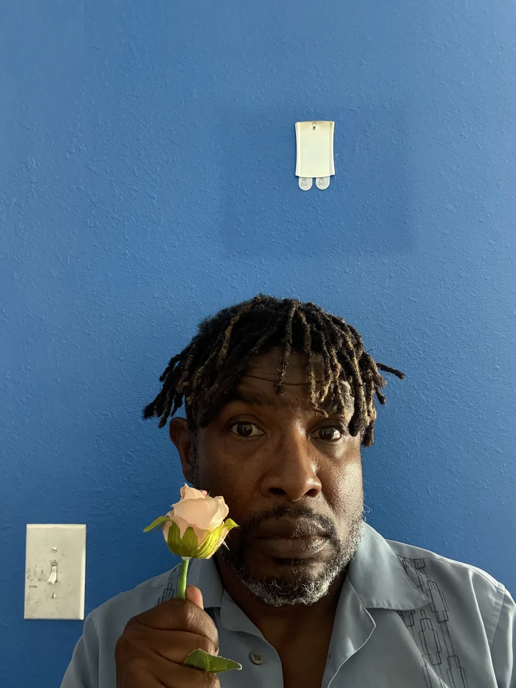 A man holds a pink rose to his face against a blue wall, in a publicity photograph for The Unforgivable Crime of Love